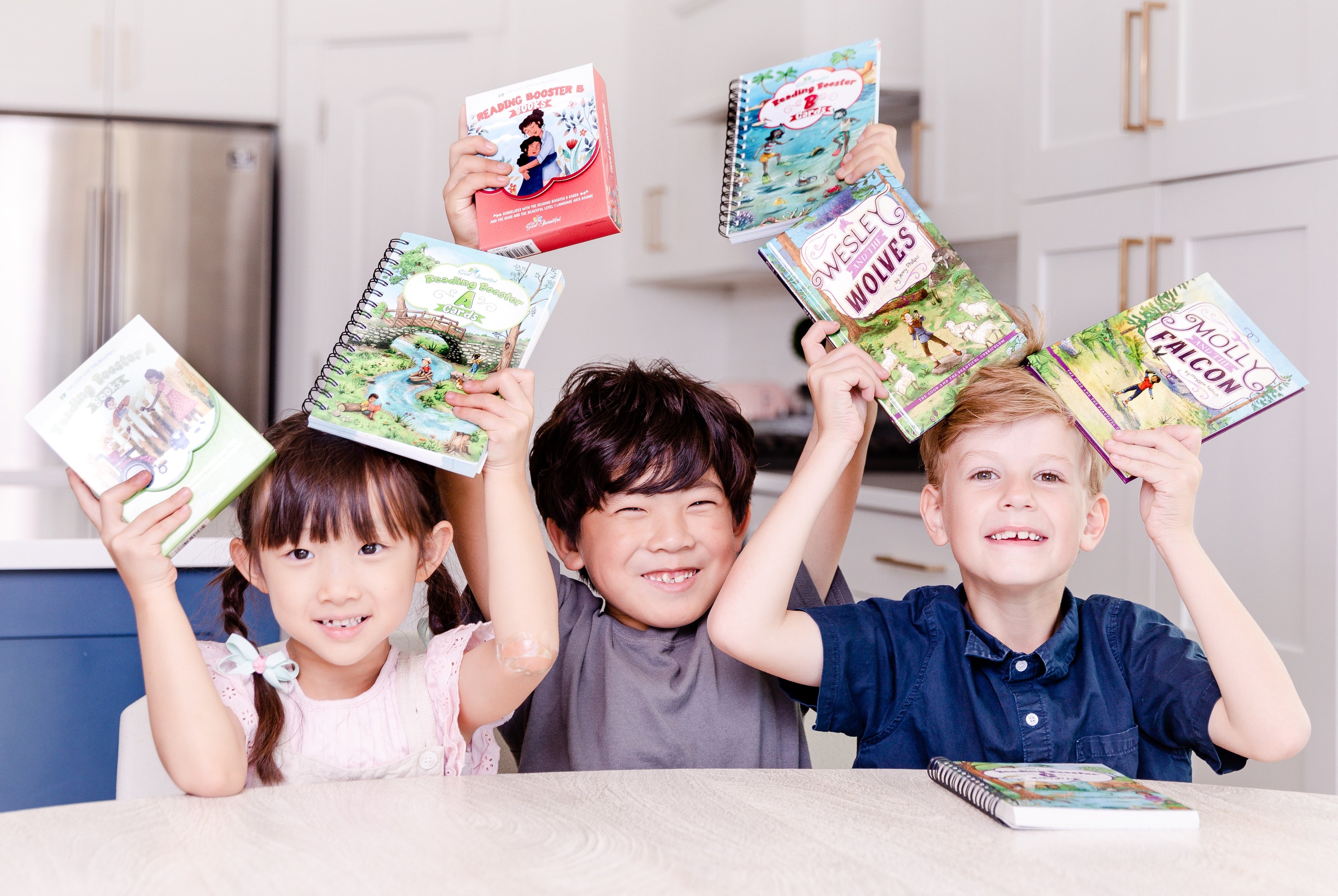 Three kids sitting at a table, smiling, and holding up their reading booster sets | The Good and the Beautiful