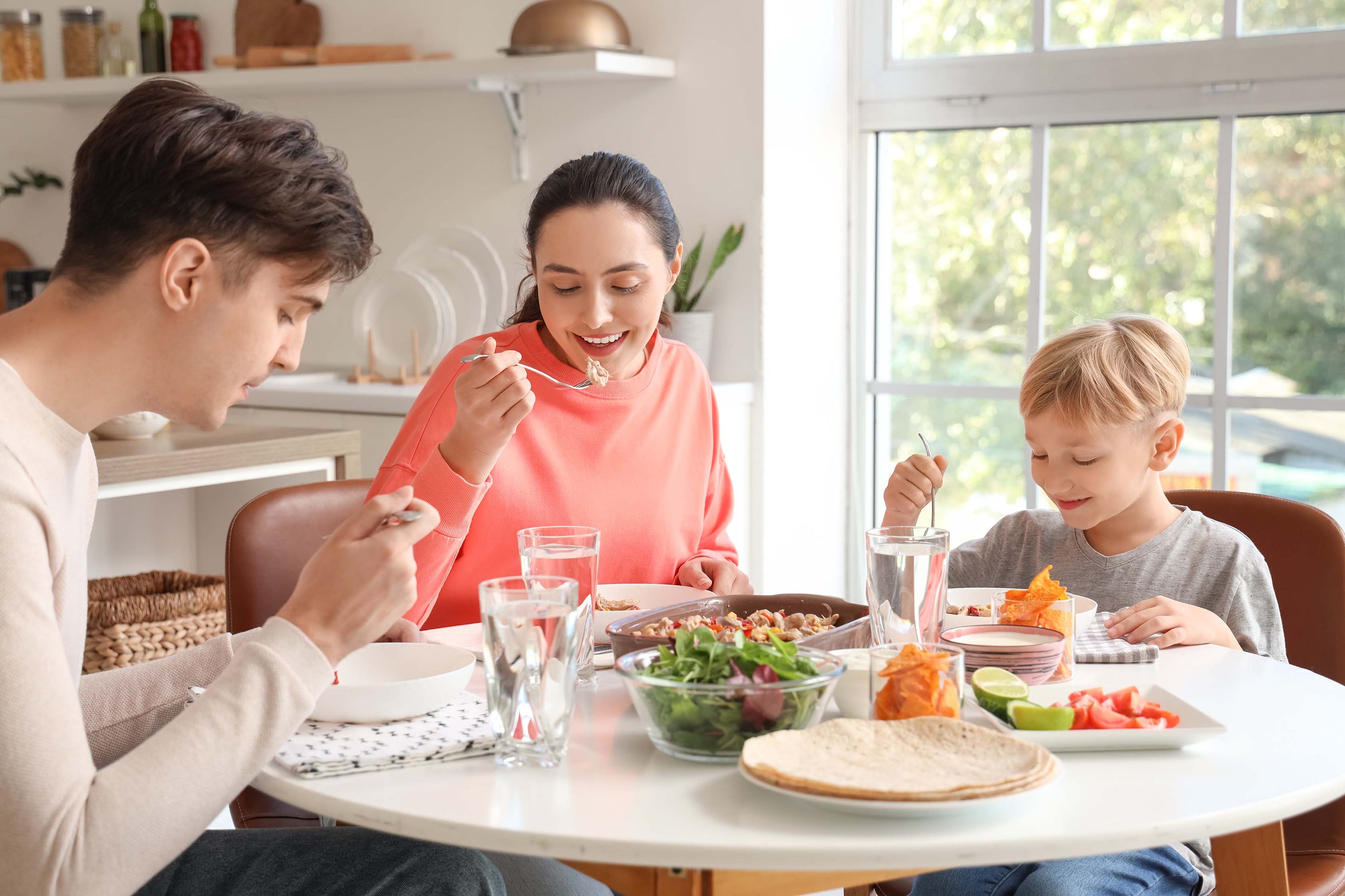 A family of three sitting at a table with dishes of food filling the table as they each eat from their own plate | The Good and the Beautiful