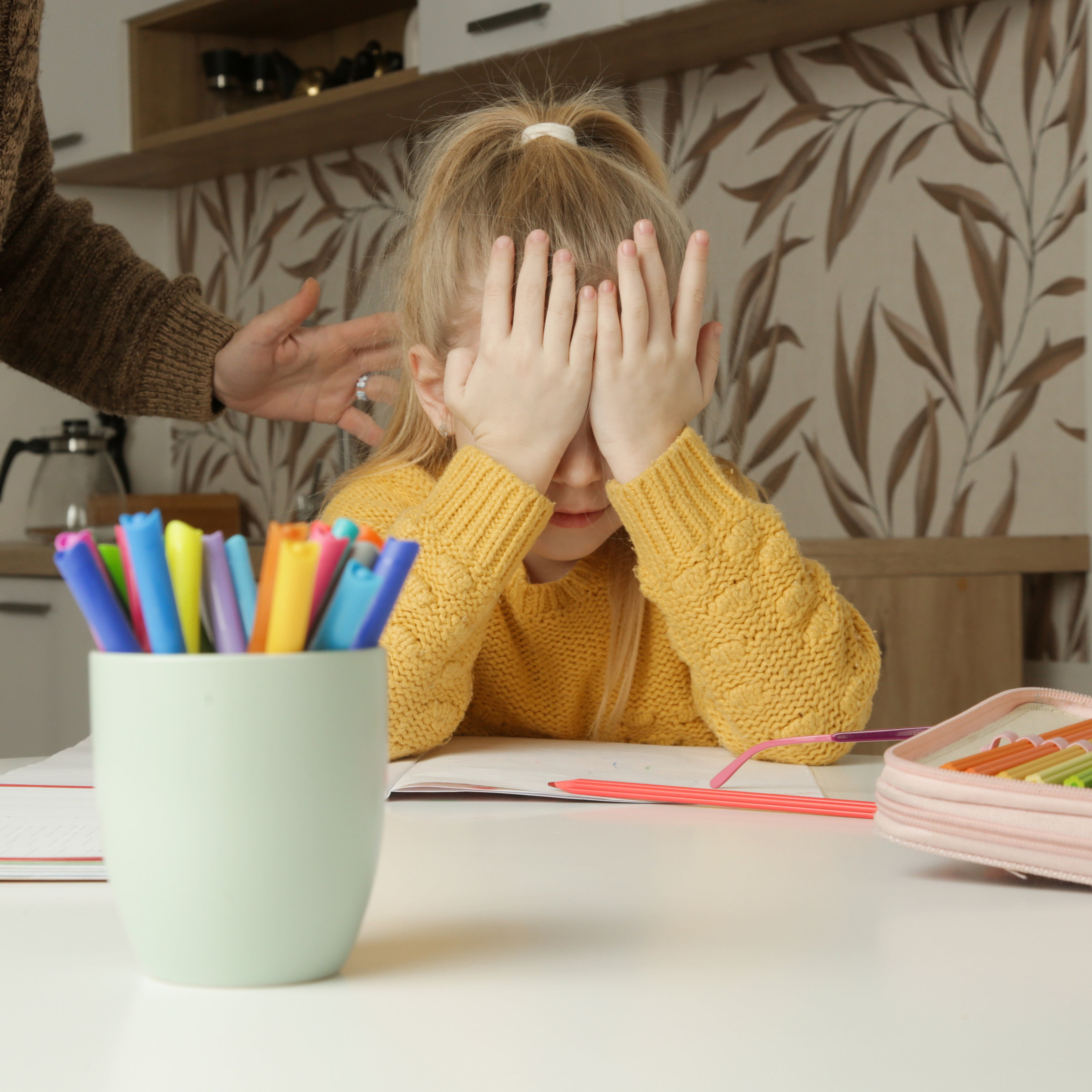 A young girl sitting at a table with her hands over her face and elbows on the table with paper and pencils in from of her | The Good and the Beautiful