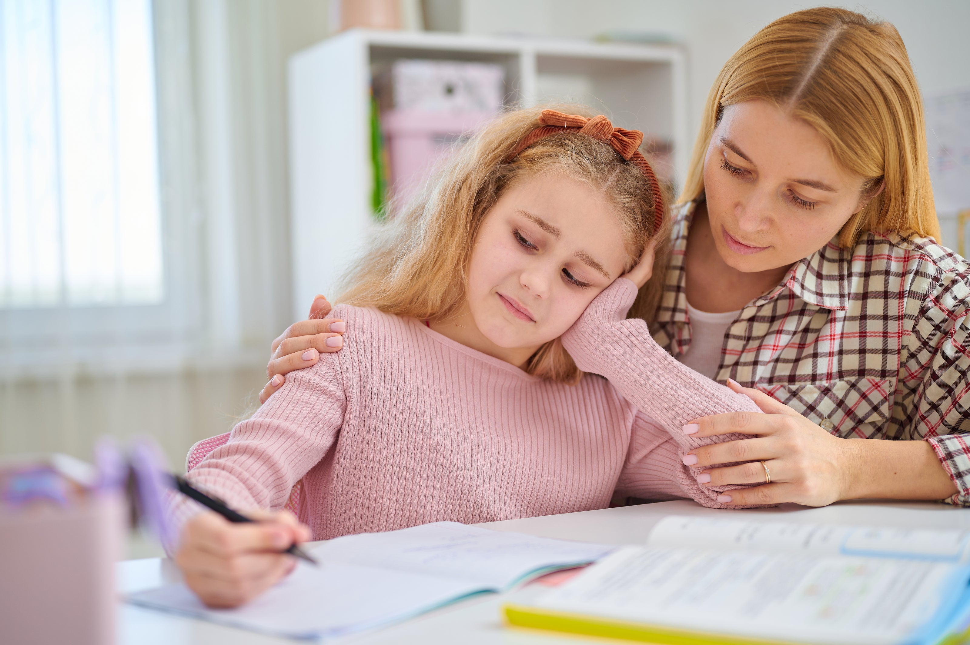 A young girl writing in a book with a sad look on her face while her mom sits behind her holding her shoulders | The Good and the Beautiful