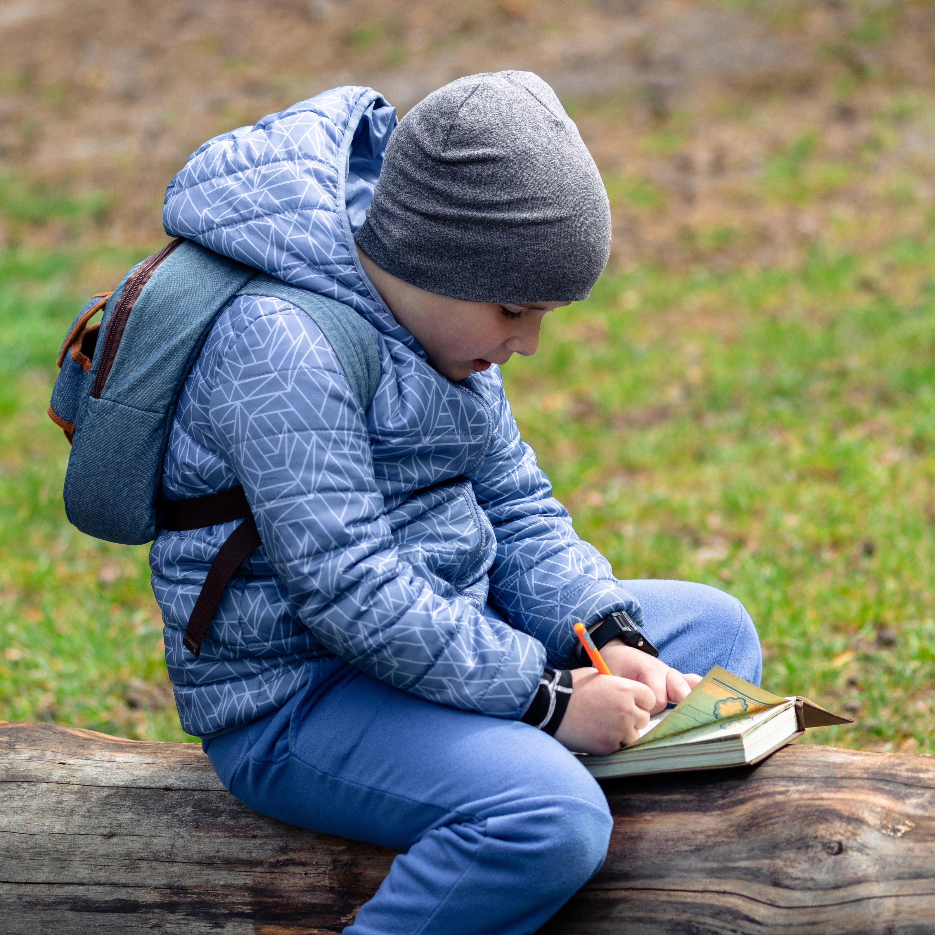 Child in blue jacket and gray beanie sitting on a log outdoors, writing in a notebook.