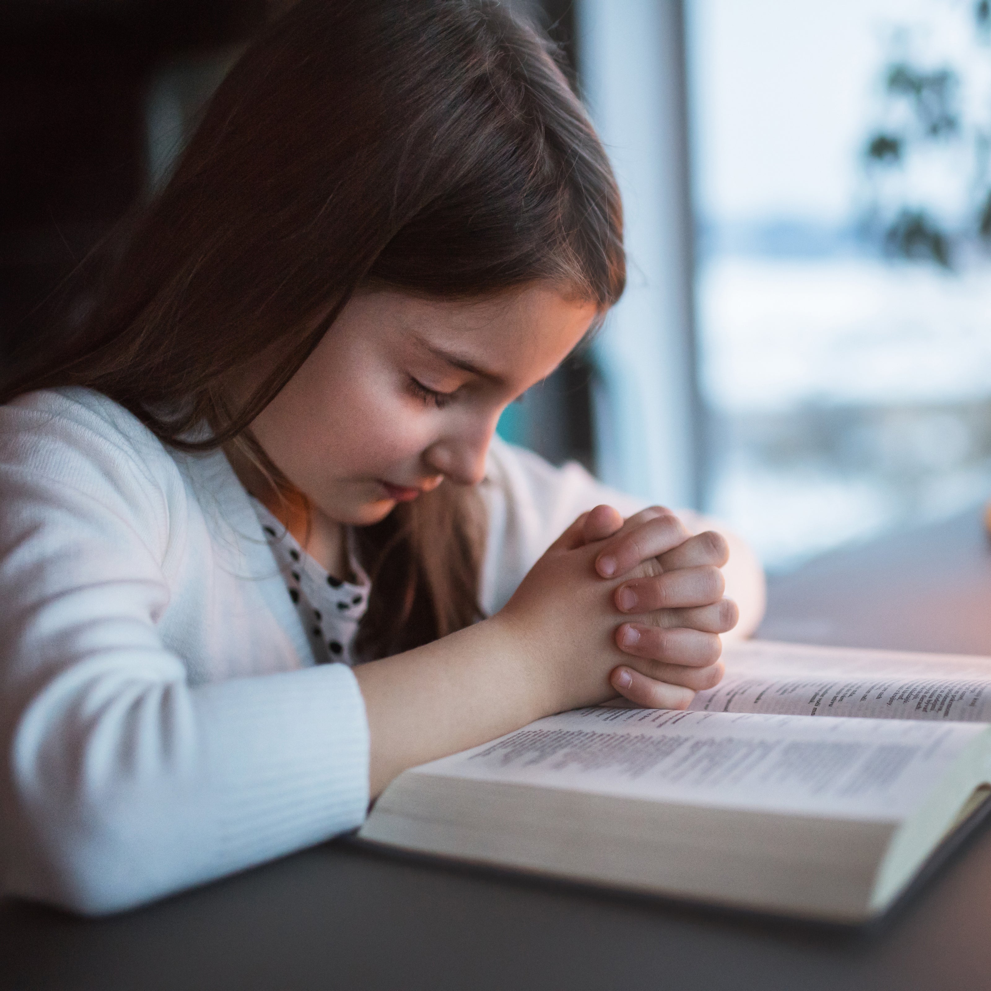 A young girl kneeling and praying with her eyes closed over an open bible | The Good and the Beautiful