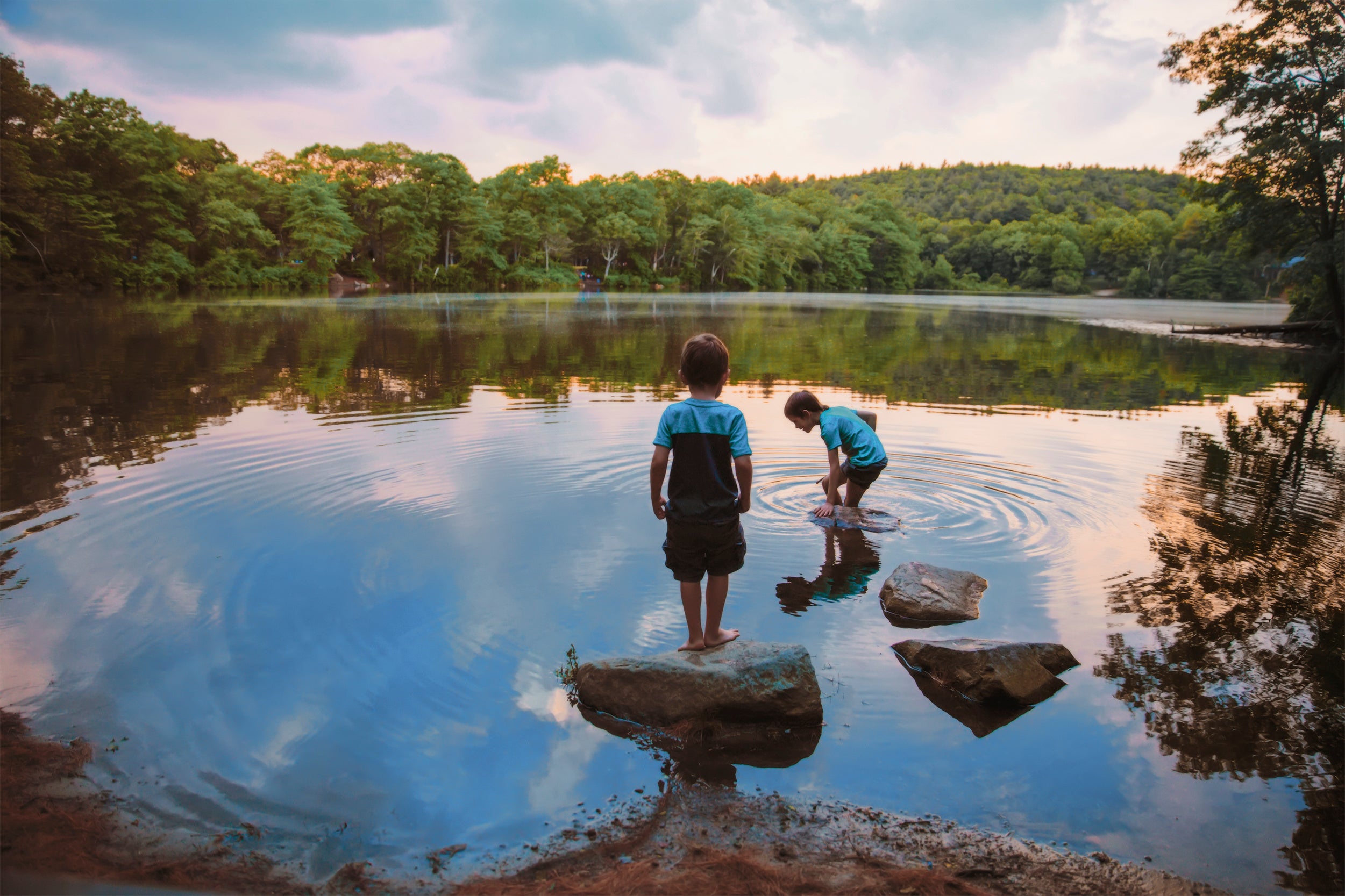 Two children playing on rocks by a lake with trees in the background