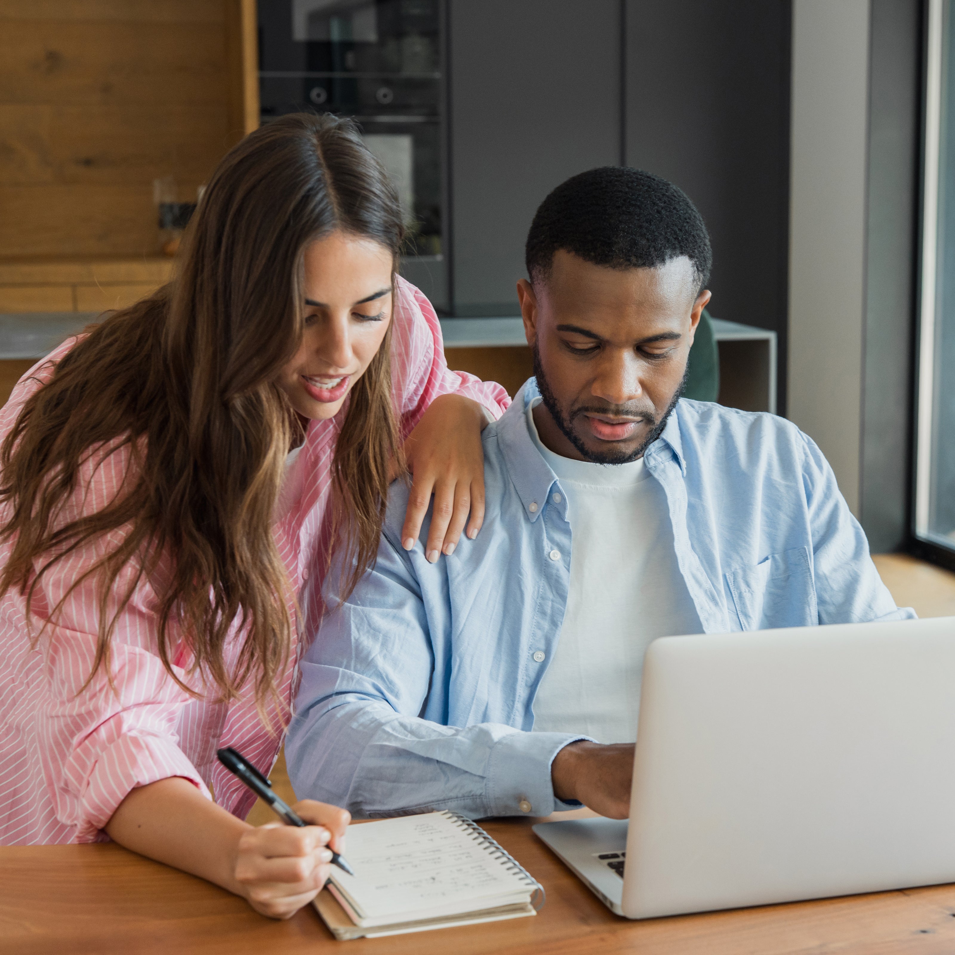 A man sitting and typing on a computer while his wife holds his shoulder and writes in a notebook | The Good and the Beautiful