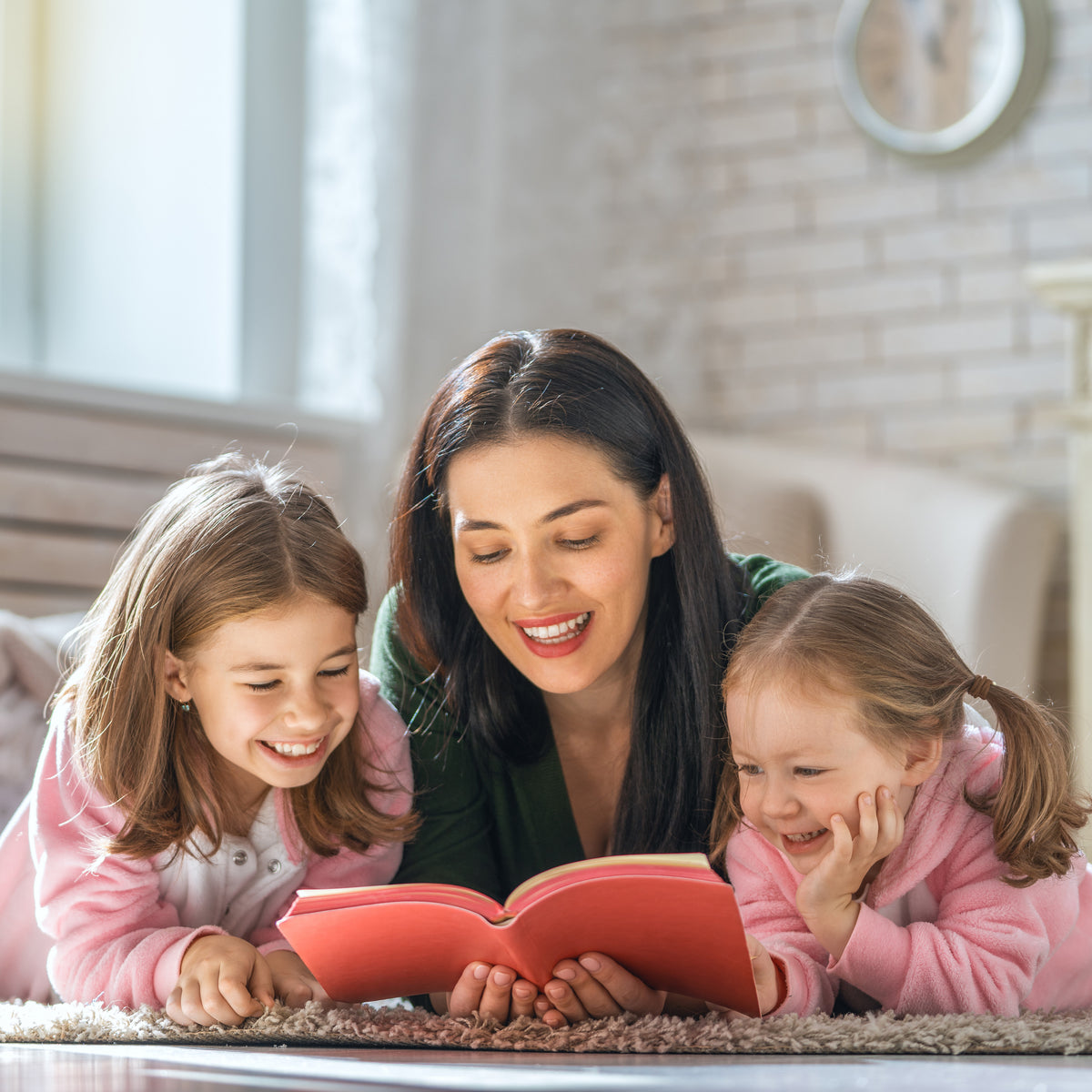 Two school-aged girls laying with their mom as they read a book together | The Good and the Beautiful