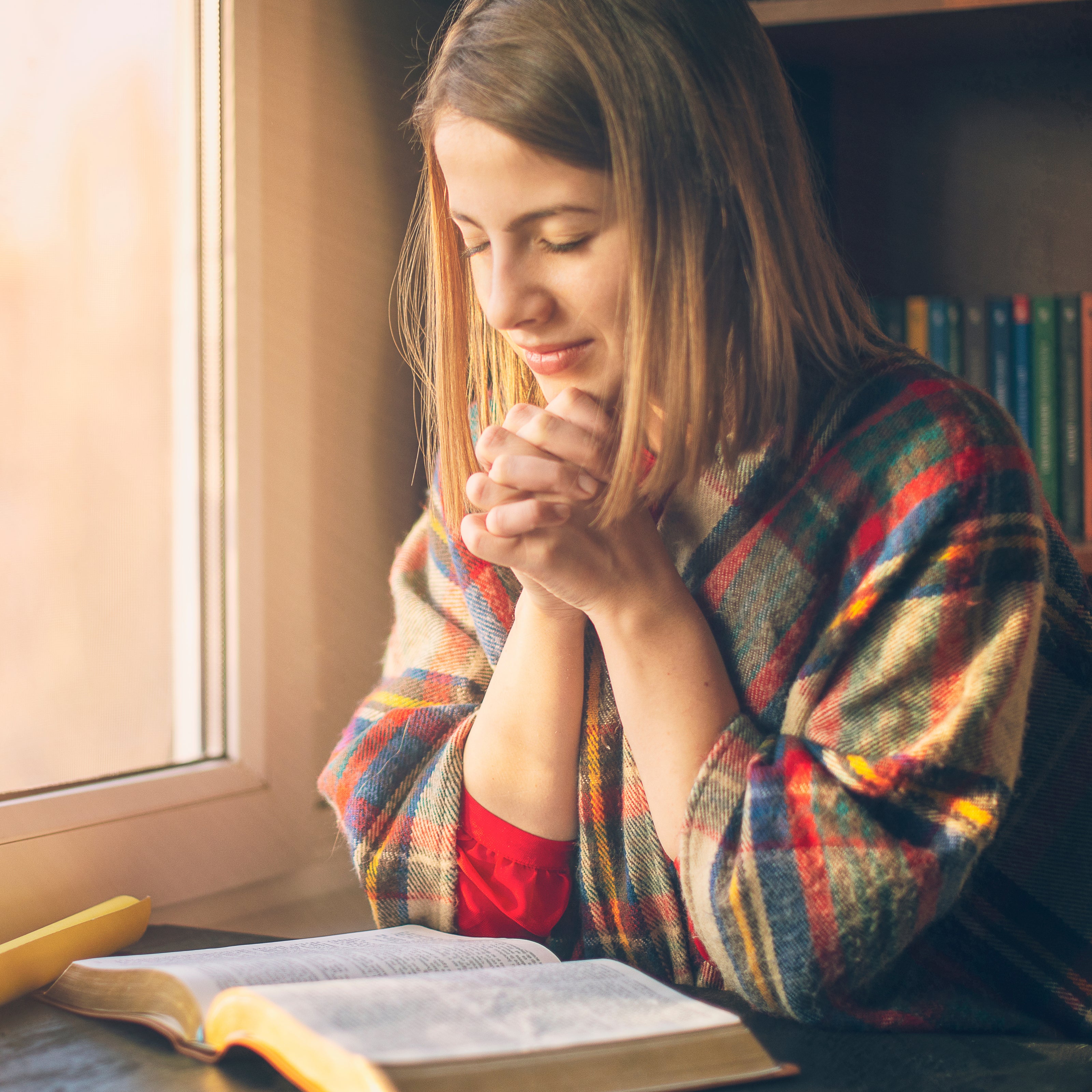 A woman praying with her elbows on the table over an open bible | The Good and the Beautiful