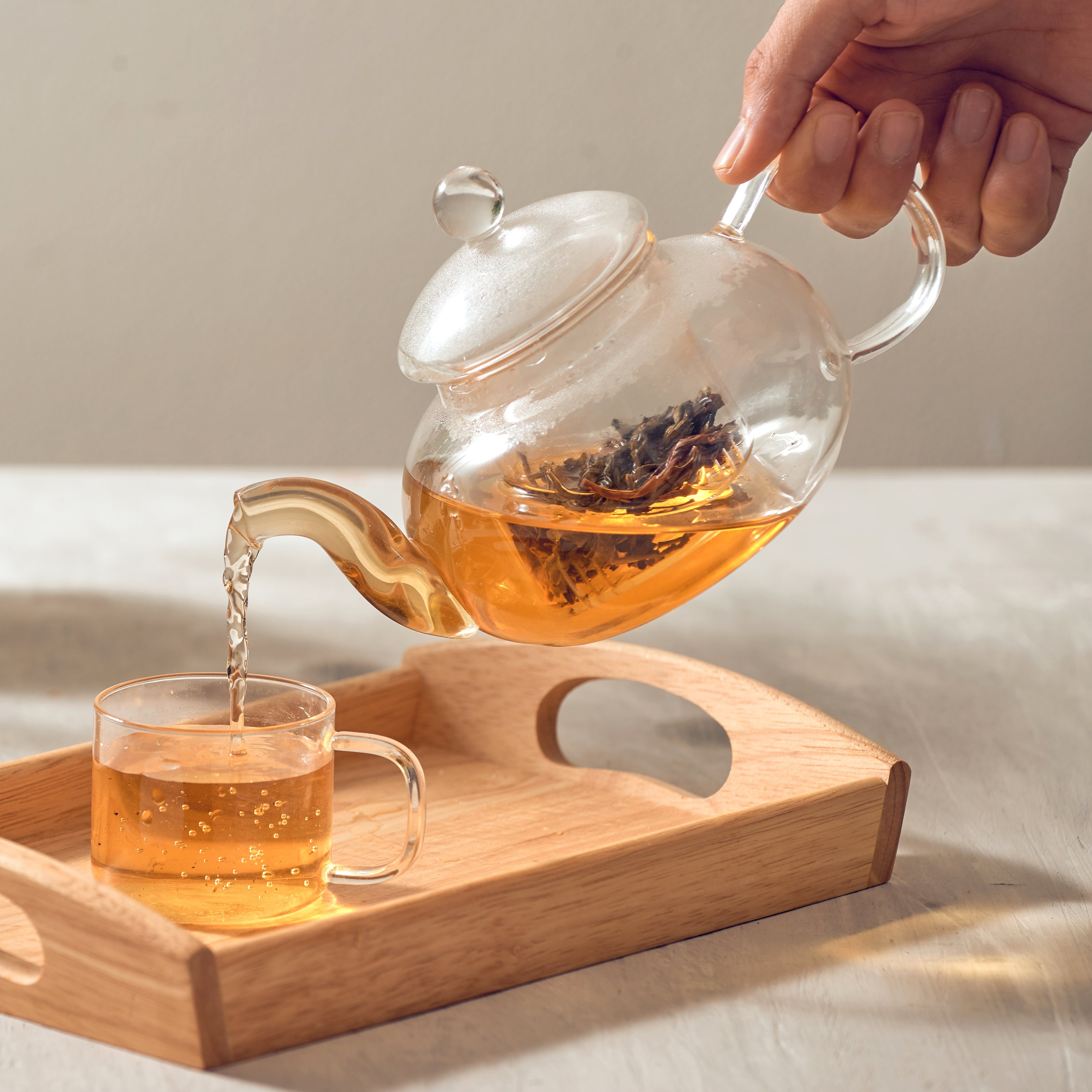 A hand pouring tea from a clear glass teapot into a clear glass teacup | The Good and the Beautiful