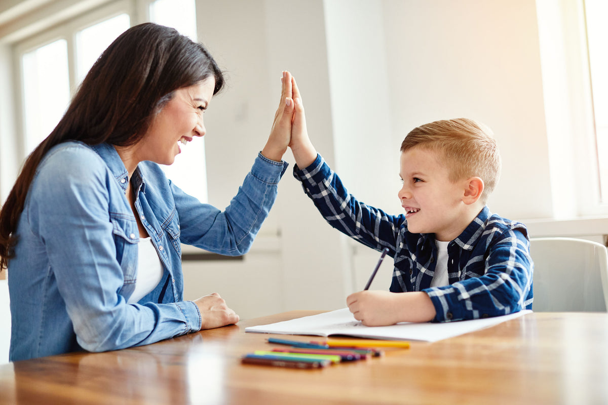 A school-aged boy writing in a notebook while giving his mom a high-five | The Good and the Beautiful