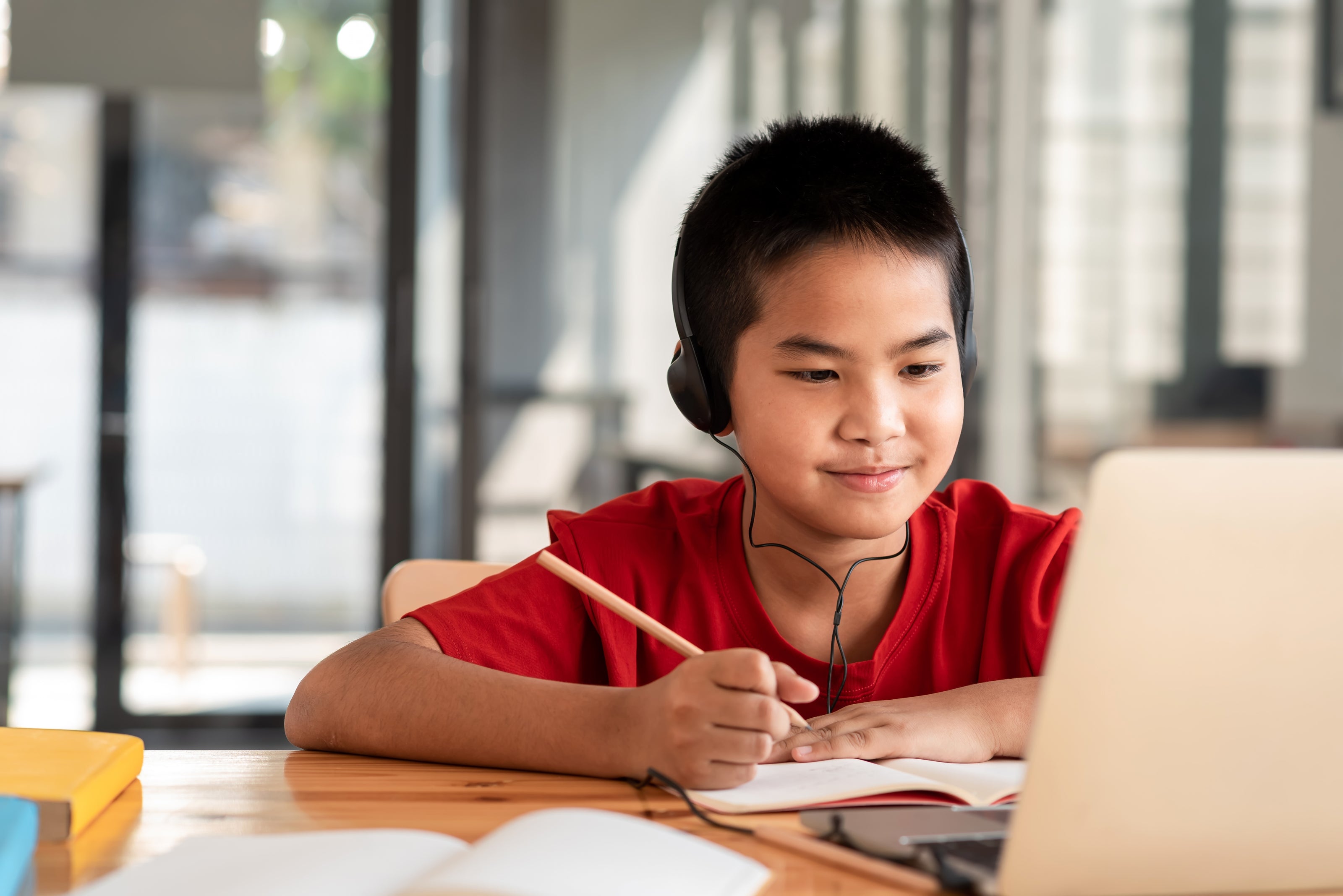 A school-aged boy sitting in front of a laptop computer with headphones on and writing with a pencil | The Good and the Beautiful