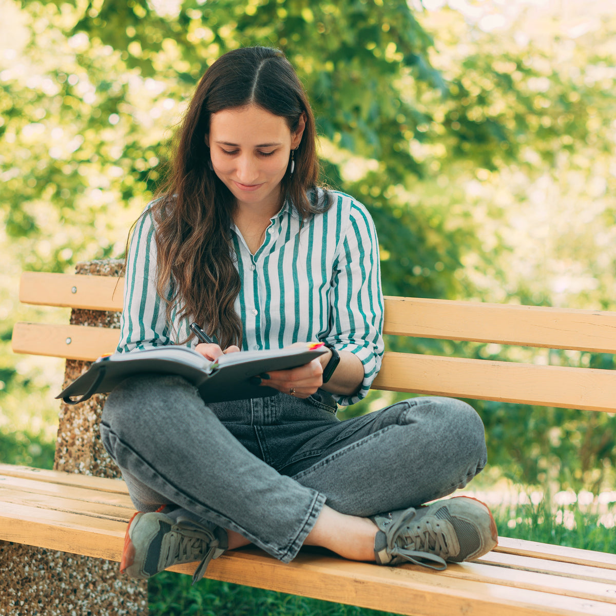 A woman sitting outside on a bench with her feet crossed as she smiles and writes in a journal | The Good and the Beautiful