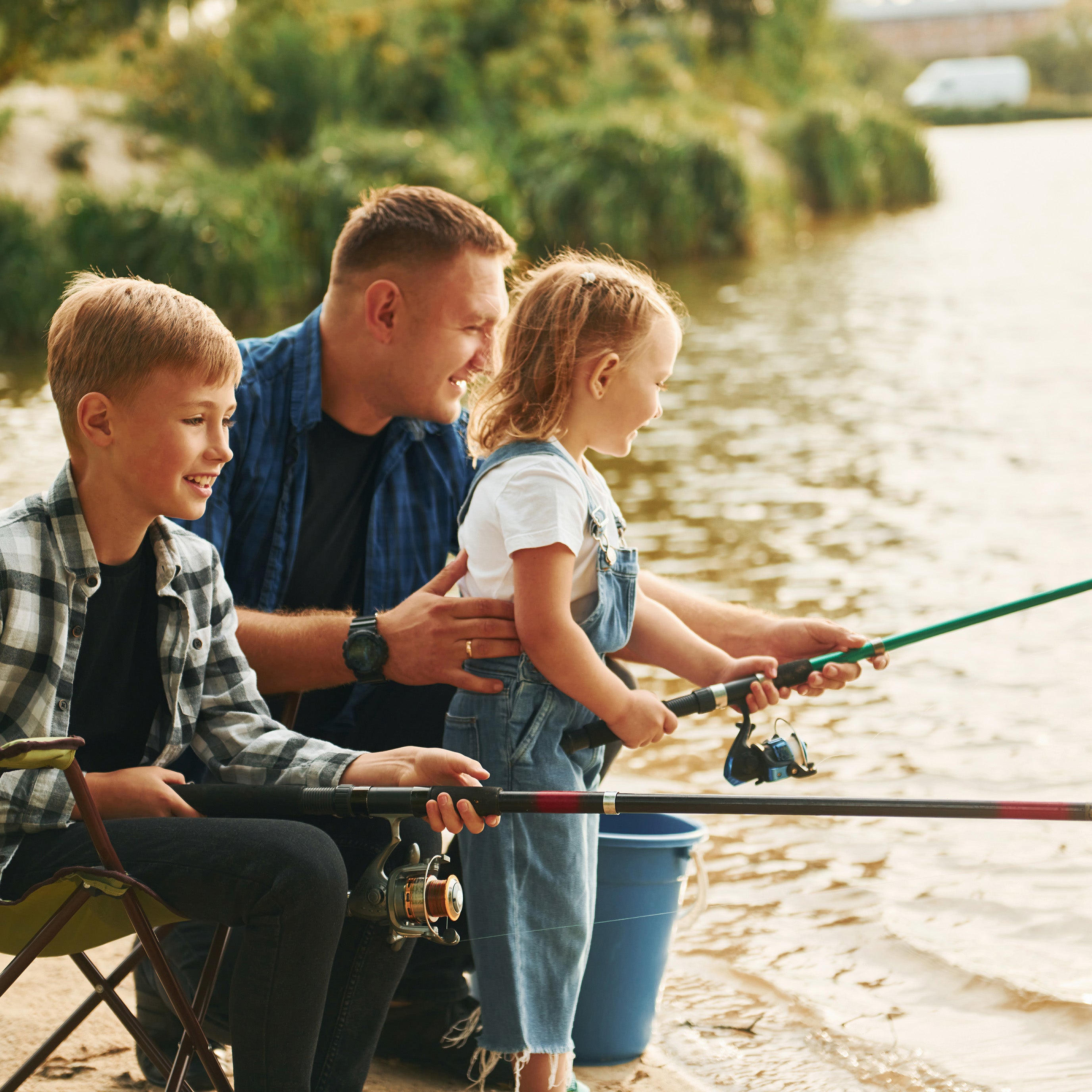 Two school-aged kids holding fishing poles by a body of water while their father sits behind them and helps | The Good and the Beautiful