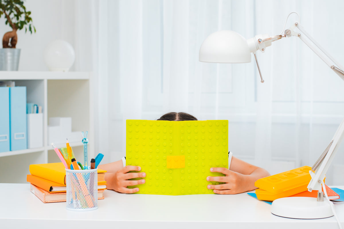 A child sitting at a desk while hiding their face behind an open notebook | The Good and the Beautiful