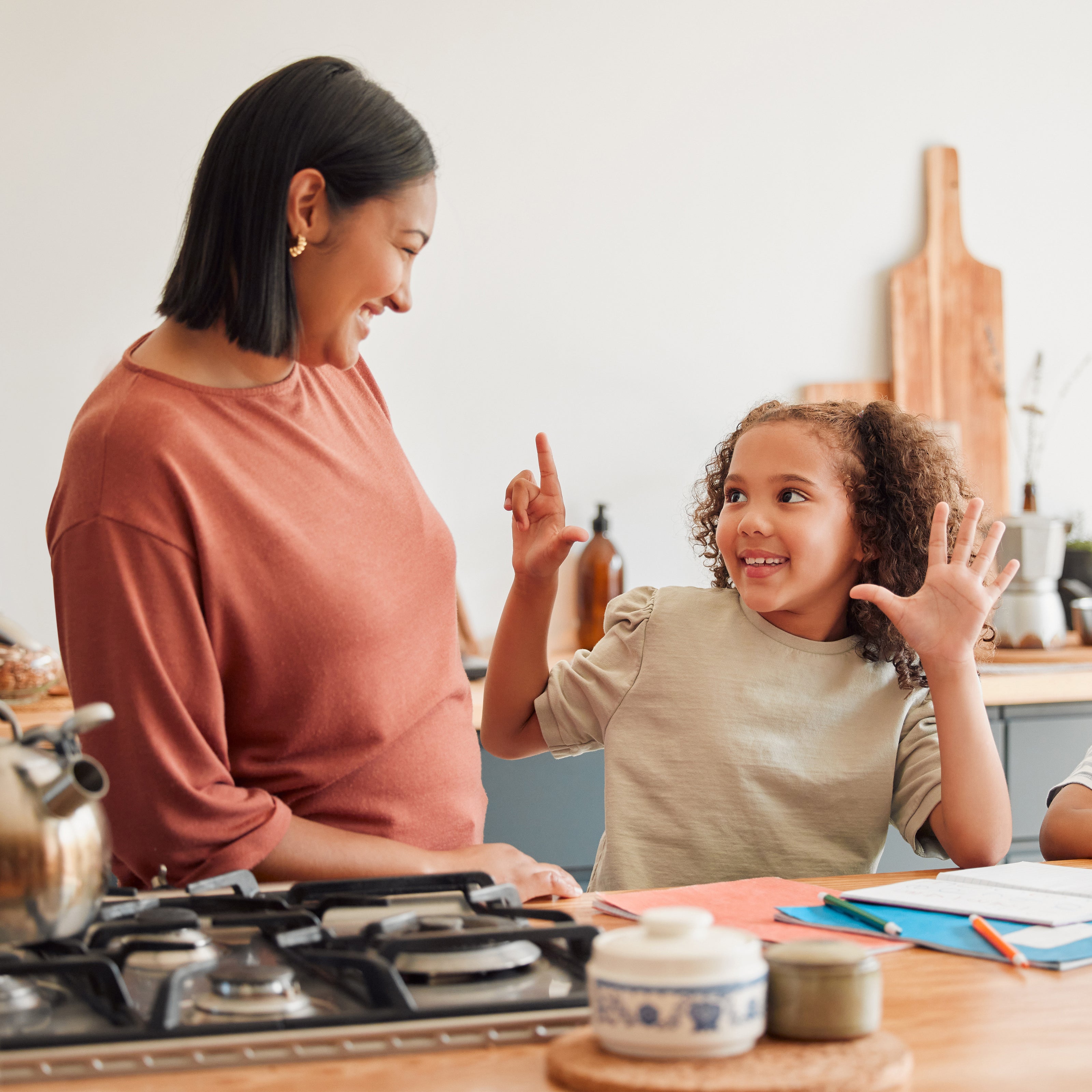 A young girl holding up seven fingers to her mom with paper and pencils in front of her and her mom smiling back at her | The Good and the Beautiful