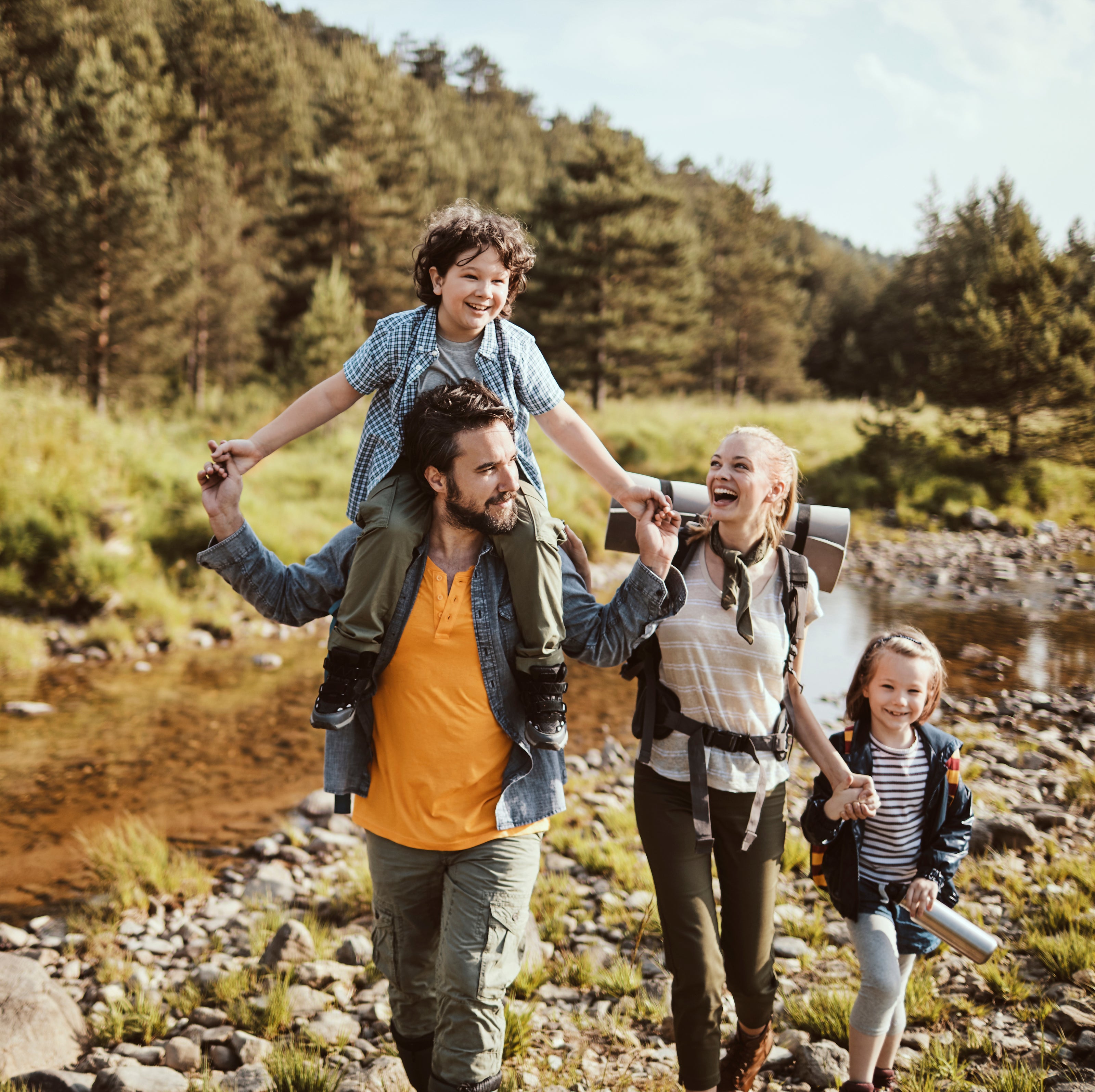 A family of four, son on his dad's shoulders and daughter holding hands with mom, are walking through a rocky area with a river and forest in the background | The Good and the Beautiful