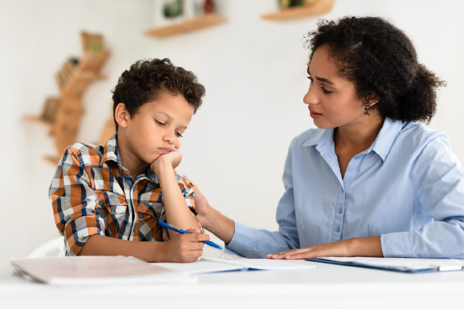 A school-aged boy writing in a notebook with his arm supporting his head and his mom looking at him while rubbing his shoulder | The Good and the Beautiful