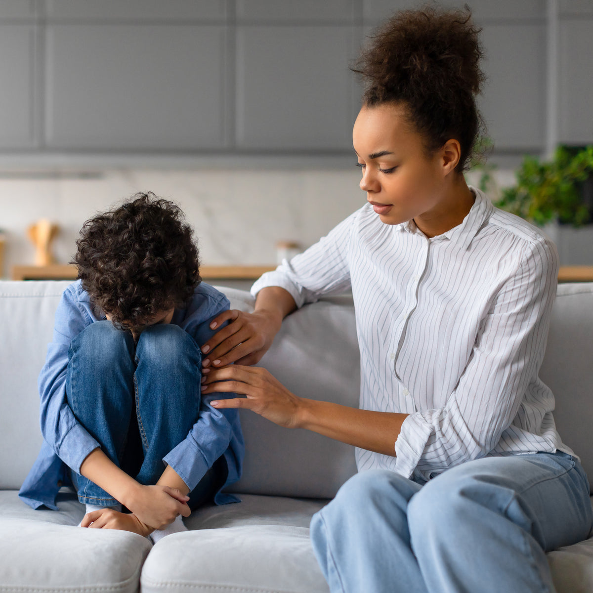 A school-aged boy sitting on a couch with his face in his knees as his mom sits next to him holding his arm | The Good and the Beautiful