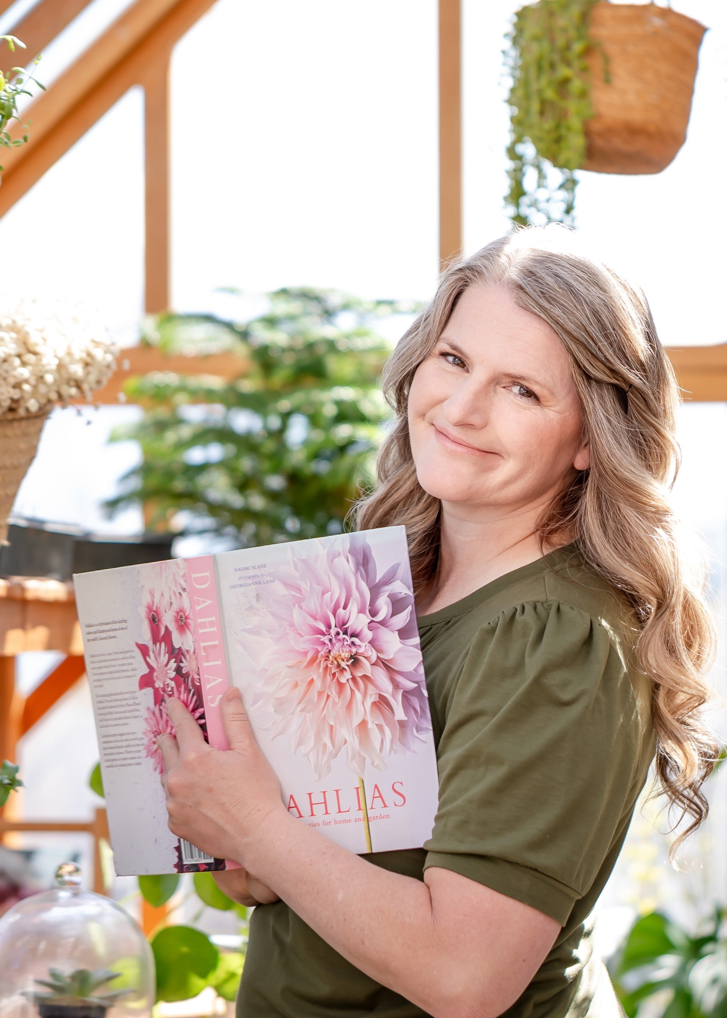 A portrait of Jenny Phillips in her sunny green house and holding a book about dahlia's | The Good and the Beautiful