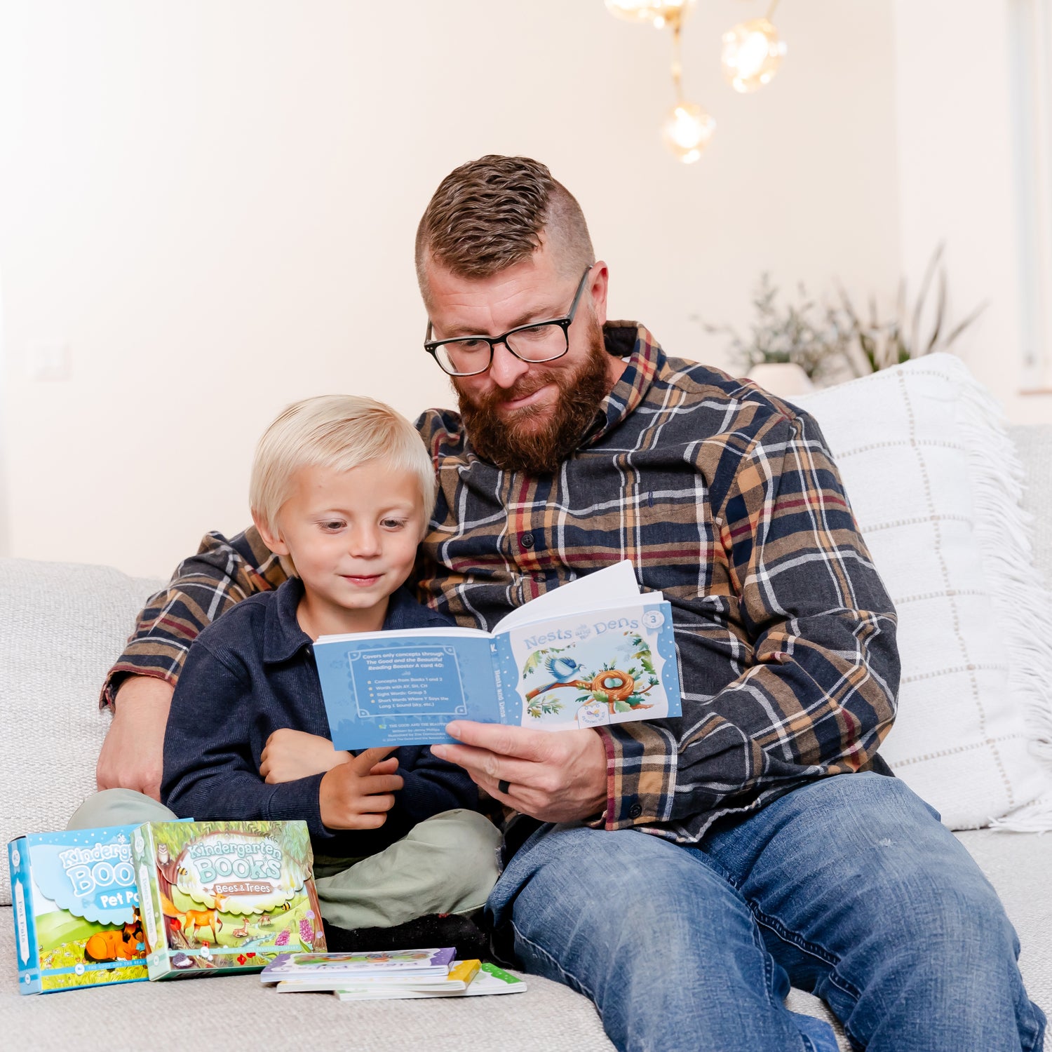 A father with his arm wrapped around his young son while he reads a book and more Kindergarten books displayed next to them | The Good and the Beautiful