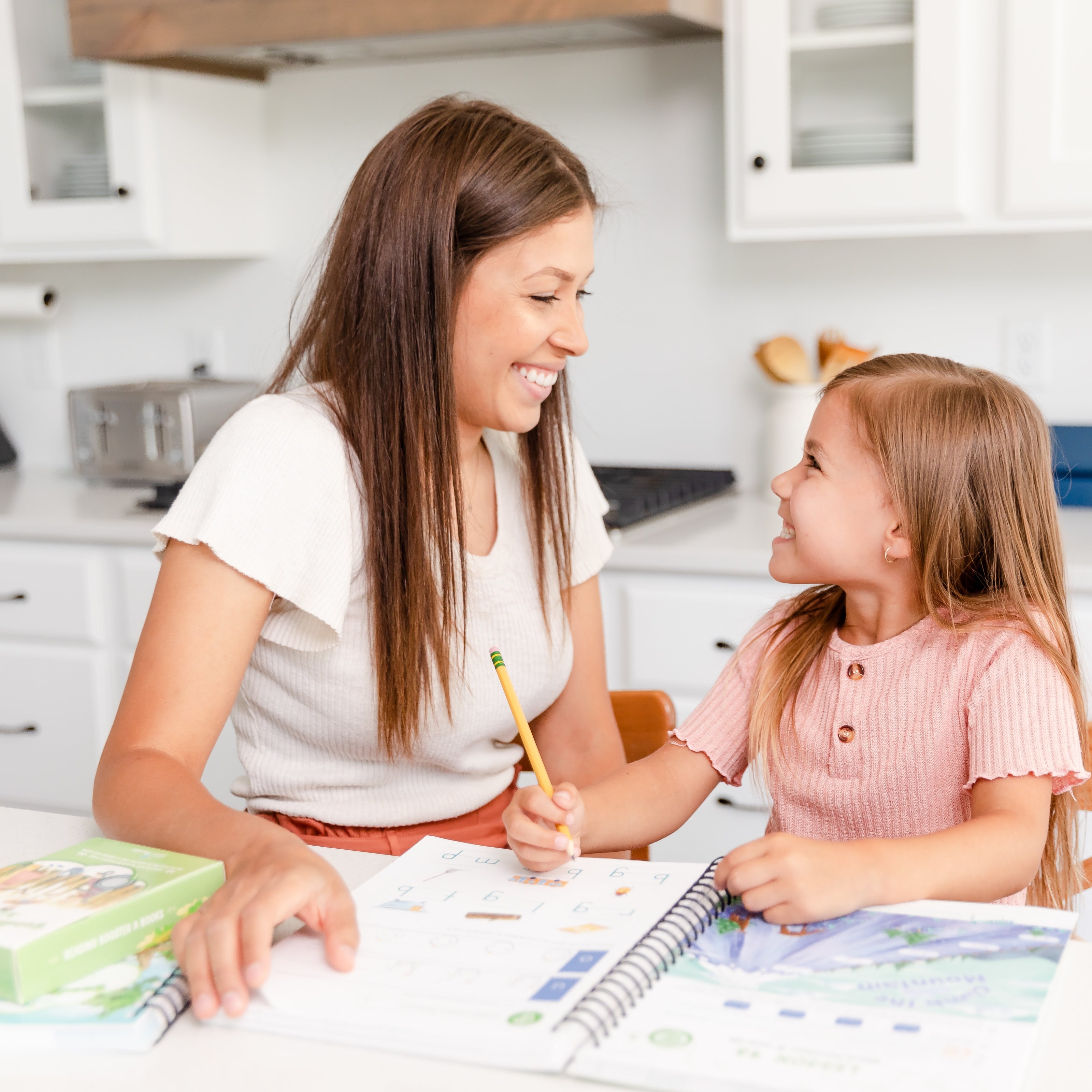 A school-aged girl smiling up at her mom as the girl writes in her language arts level K course book | The Good and the Beautiful