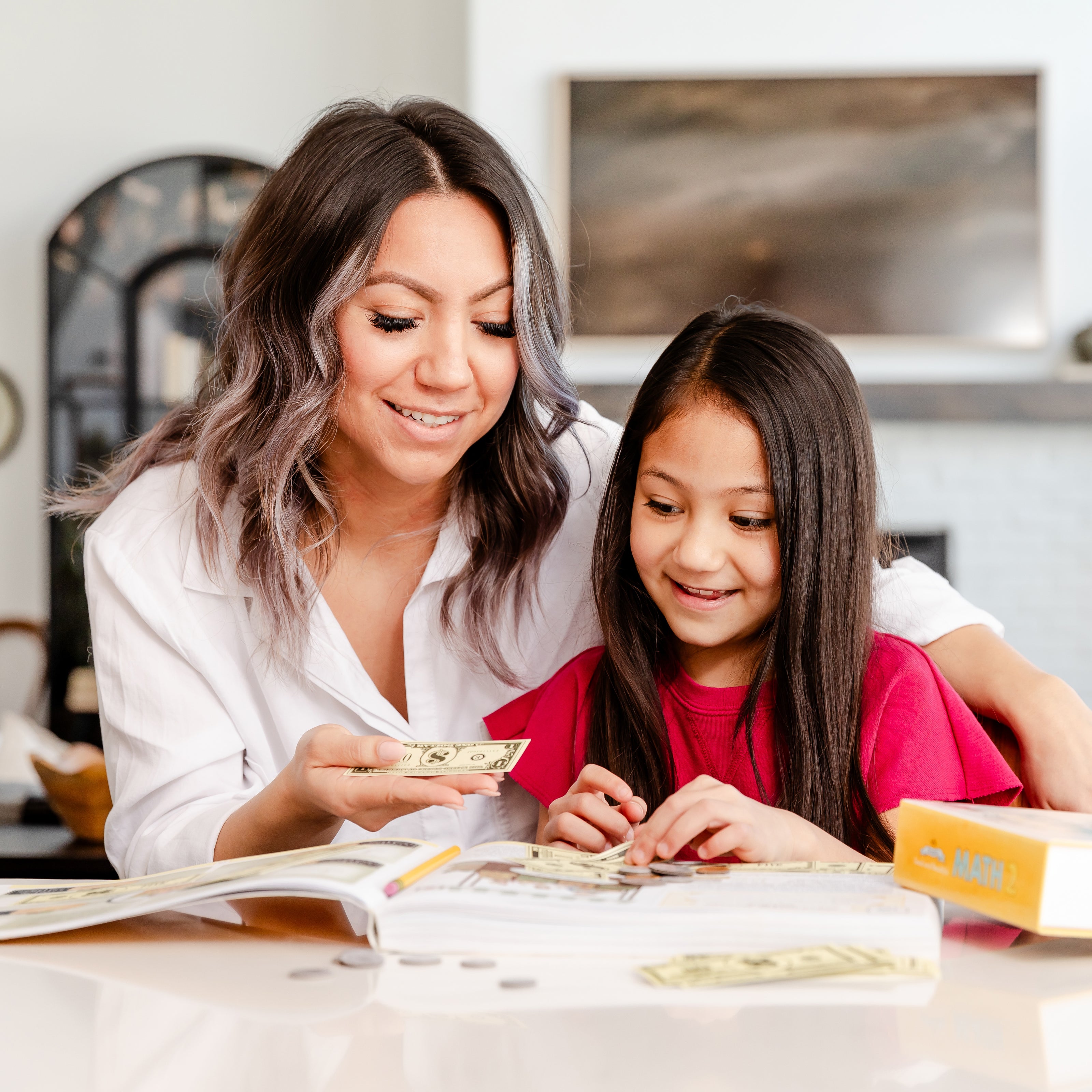 A mother and her young daughter sitting at a table as the mother holds a pretend dollar and helps her daughter with the math 2 curriculum | The Good and the Beautiful
