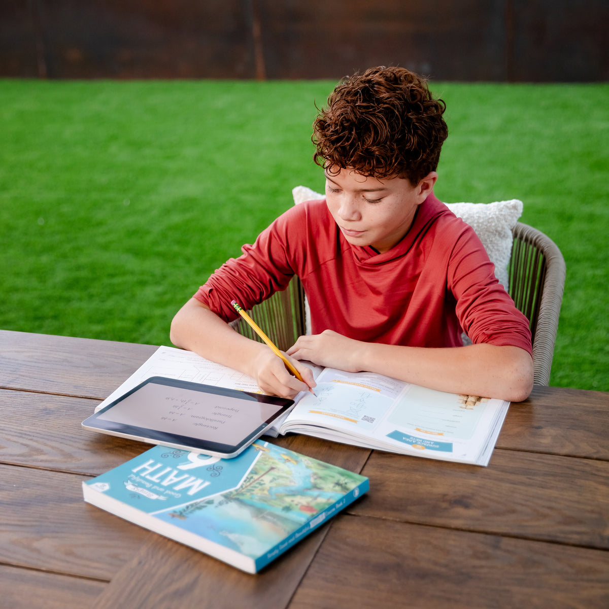 A school-aged boy sitting at a table outside and working in a level six math course book with a tablet and course book | The Good and the Beautiful