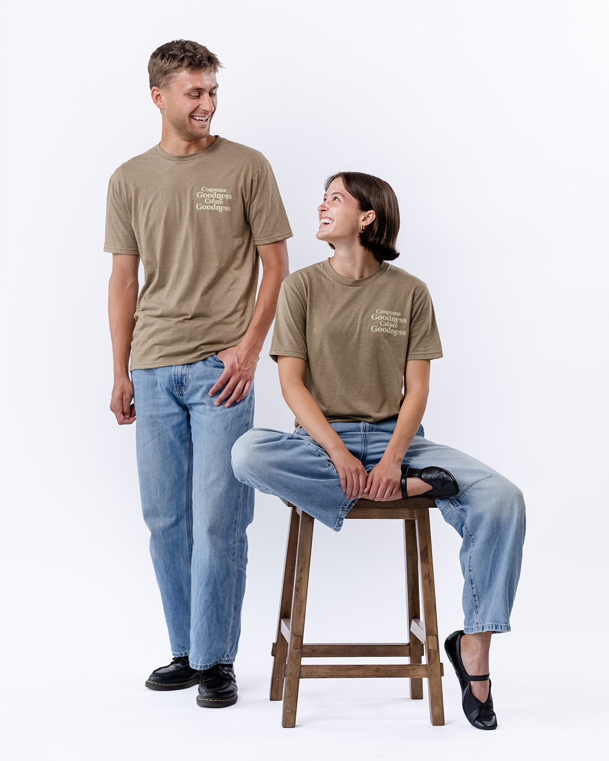 A man standing next to a woman who is sitting on a stool as they pose with brown t-shirts that say "Consume Goodness Create Goodness" | The Good and the Beautiful
