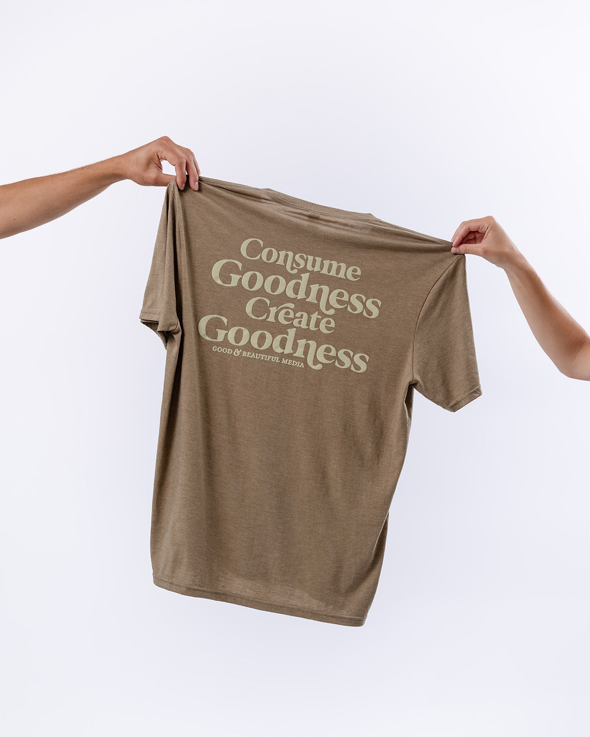 Two arms holding up a brown t-shirt with the text "Consume Goodness Create Goodness" | The Good and the Beautiful