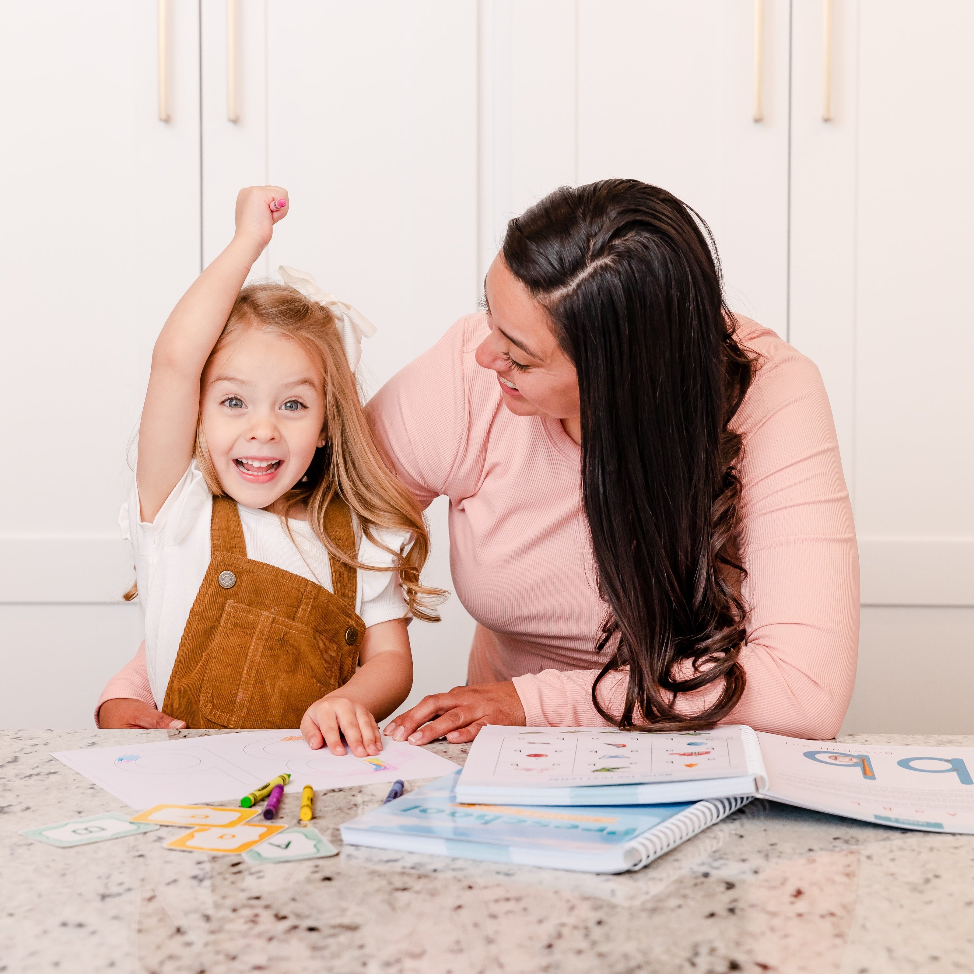 A preschool-aged girl cheering as she colors a letter page with her mother next to her | The Good and the Beautiful