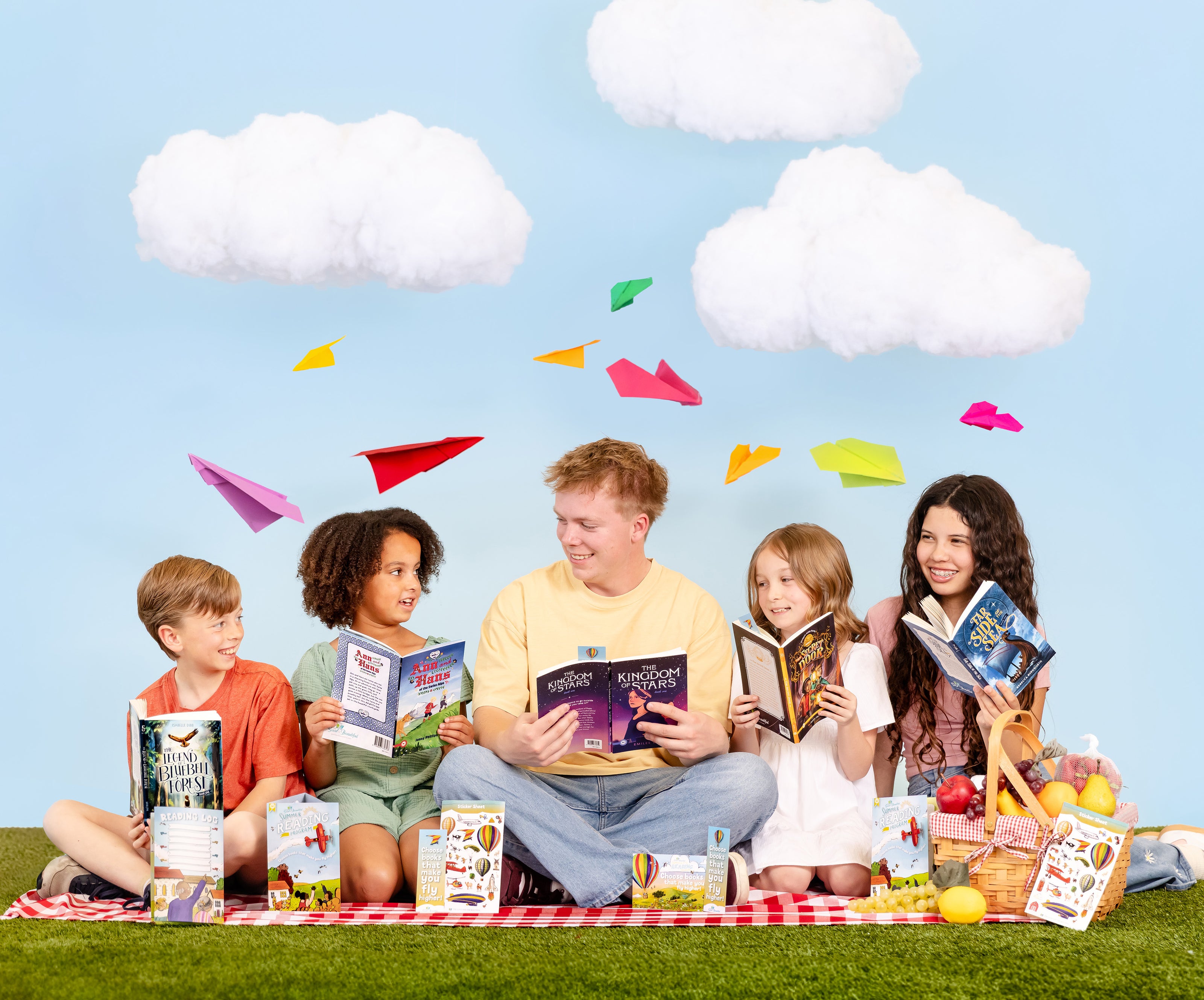 Family with children sitting on a blanket outdoors, reading books with colorful paper airplanes around them | The Good and the Beautiful 2026 Summer Reading Program