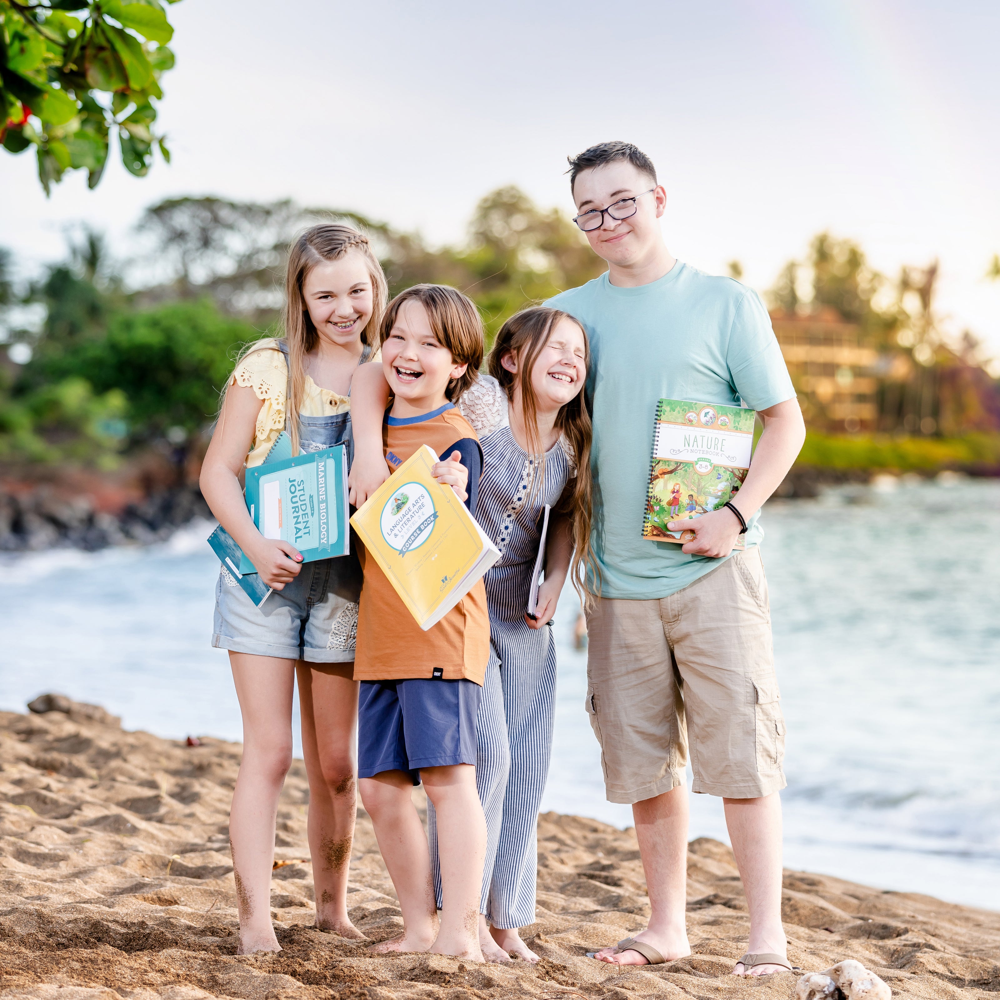 Four children standing on a beach holding The Good and the Beautiful books, with a scenic background of water and greenery.