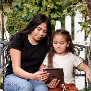 Photograph of two girls playing games on the tablet outside under the trees close shot